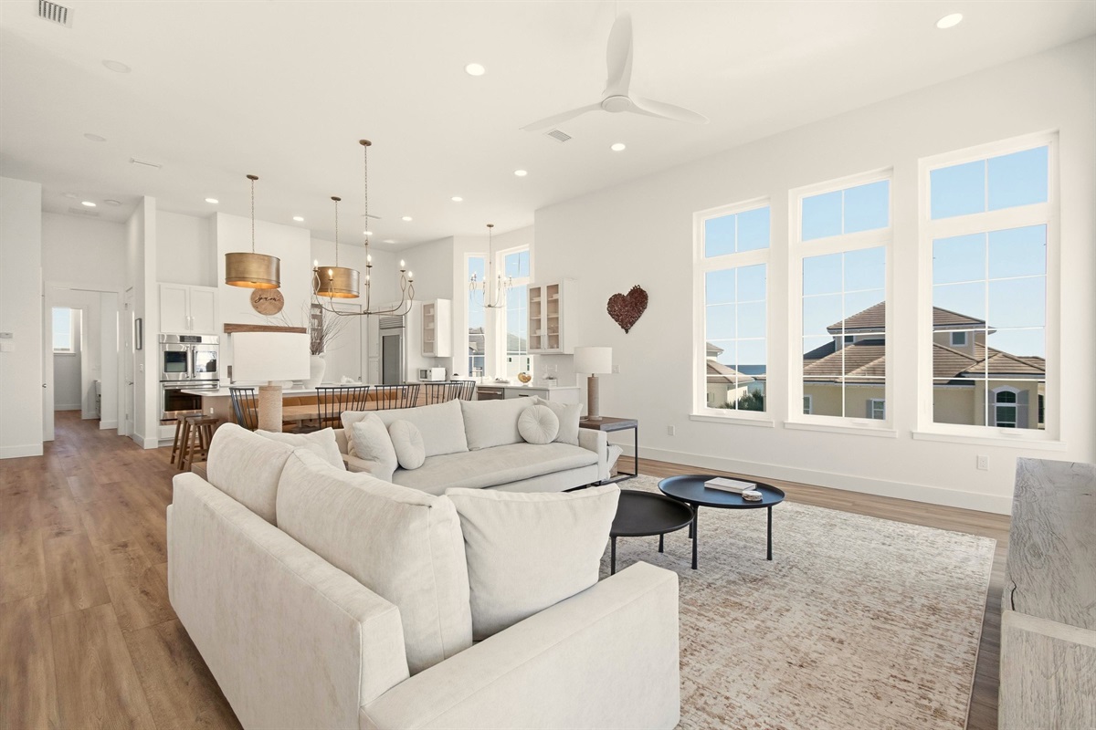 Step into a sunlit great room where tall windows frame a glimpse of the ocean, couches invite long chats, and the open kitchen keeps everyone together. Pour a drink, cue a playlist, and let the day slow down.
