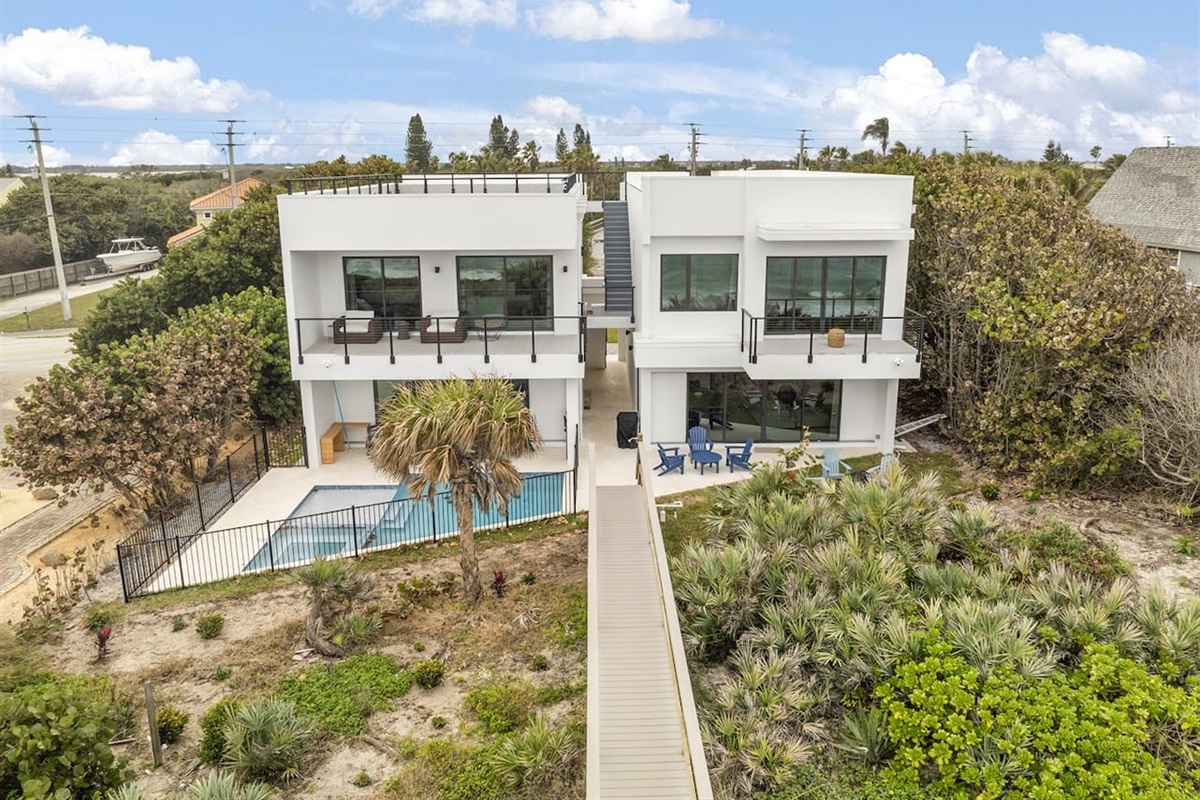 Captured by drone, this outdoor space features a beautifully designed swimming pool, surrounded by lush trees and greenery, with a charming bridge leading to the sea, creating a serene and inviting ambiance for relaxation.