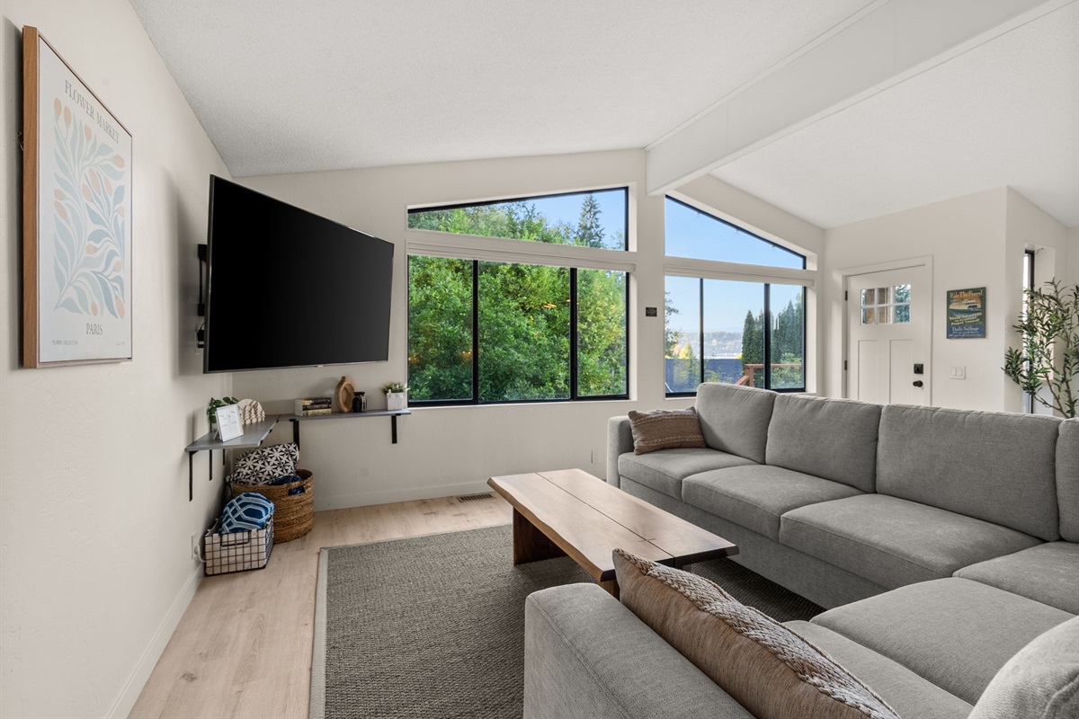 Relax in the bright, open living space featuring a comfy sectional, a large TV, and expansive windows—including unique trapezoidal panes—that bring the outdoors in