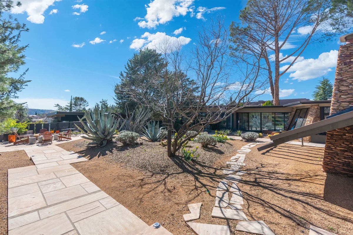 Beautifully landscaped desert courtyard with sculptural agave, winding stone pathways, and inviting lounge areas — all surrounding a stunning mid-century home with expansive glass and seamless indoor-outdoor living.