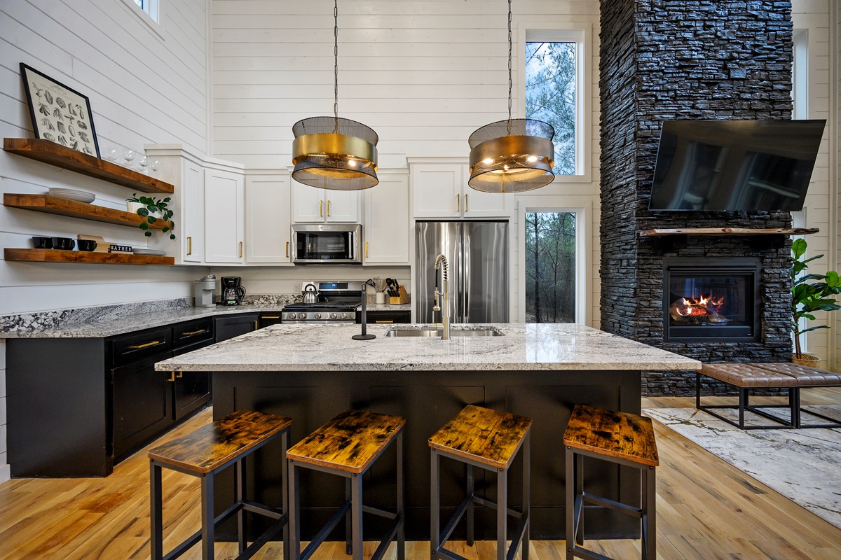 An open-concept space bringing the kitchen and living area together seamlessly — featuring a beautifully styled kitchen island, modern finishes, and a cozy glimpse of the fireplace and TV that adds warmth and inviting cabin charm throughout the space.