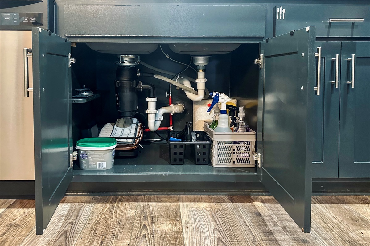 Under-sink kitchen cabinet neatly arranged with cleaning supplies, dish drying rack and mat, paper towels, and dishwashing detergent.