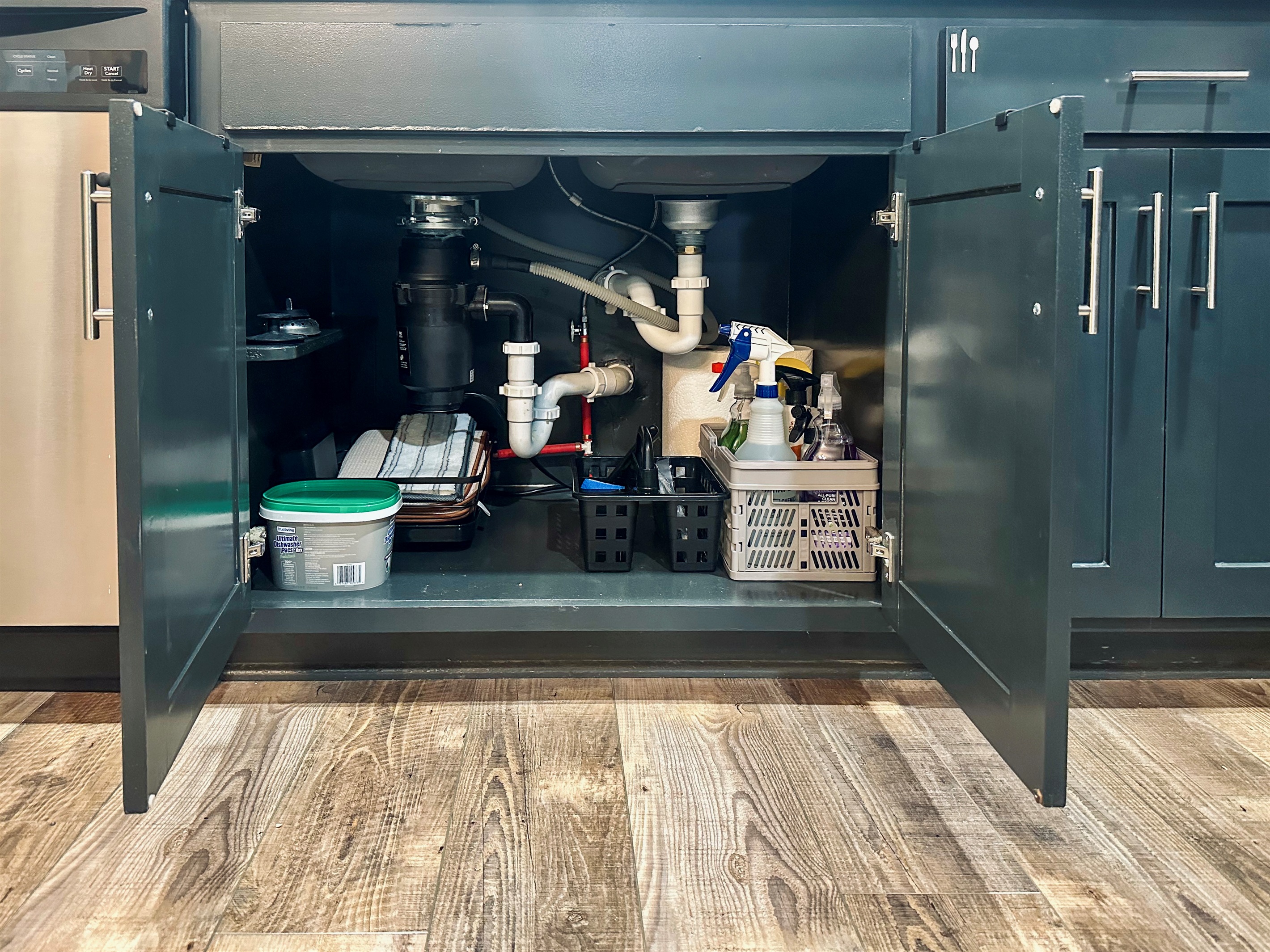 Under-sink kitchen cabinet neatly arranged with cleaning supplies, dish drying rack and mat, paper towels, and dishwashing detergent.