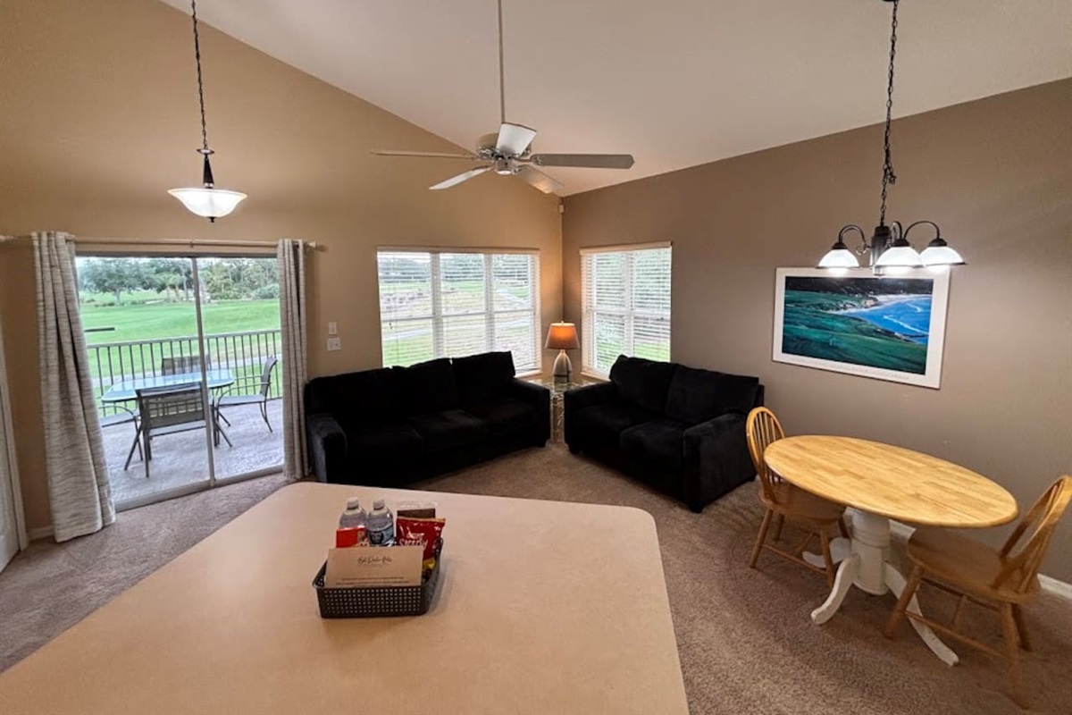 Bright living and dining space with cozy seating, vaulted ceilings, and peaceful views from the balcony doors.
