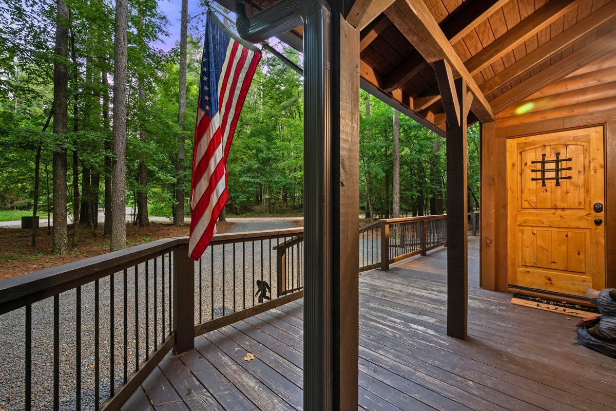 Wraparound porch with railing, American flag, and forest views — perfect for morning coffee or evening chats.
