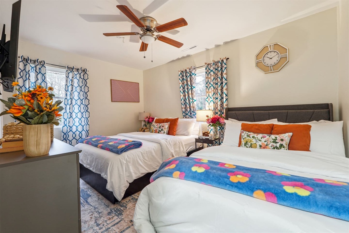 Step into a comfortable bedroom featuring two Queen beds, a ceiling fan, a TV, a clock, and a basket of fresh towels, providing everything you need for a relaxing and restful stay.

