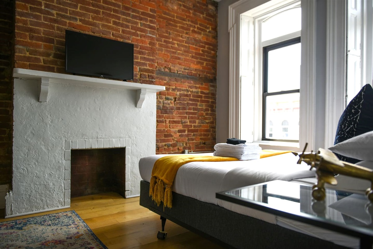 Relax in this bedroom featuring a white bed, small TV, and exposed brick walls for a modern touch. Wake up to natural light from the wide window, creating a bright and cozy ambiance. The bedside lamp sets the perfect mood for a peaceful night’s rest.