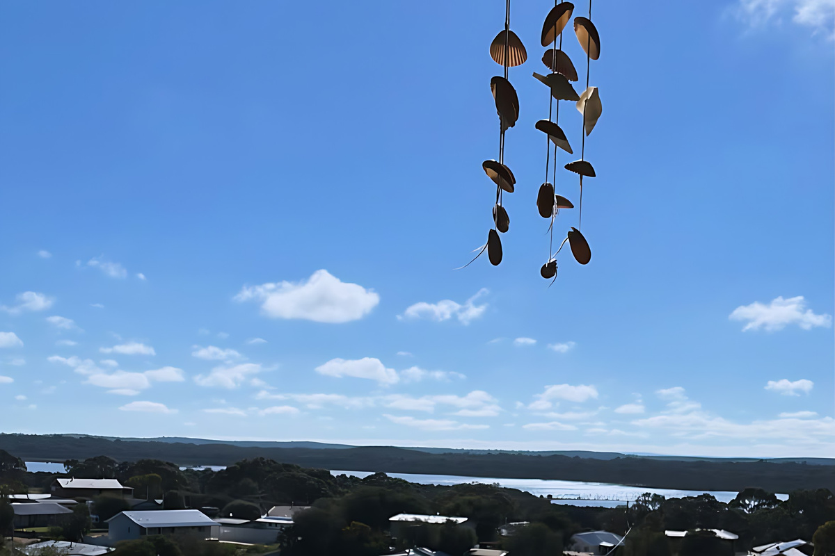 A shell wind chime hangs from the veranda eave, with Lake Preston and the Yalgorup National Park shoreline visible beyond the rooftops of Preston Beach on a clear day.