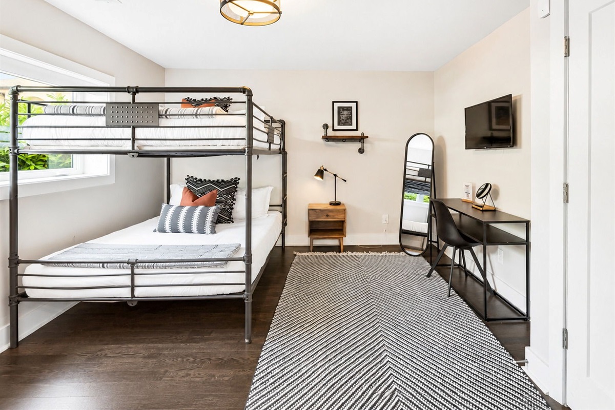 Metal bunk bed with striped bedding offers a queen over queen setup - Includes a wood nightstand, reading lamp, and minimalist desk with modern mirror - Bright window and geometric rug enhance the clean, contemporary vibe