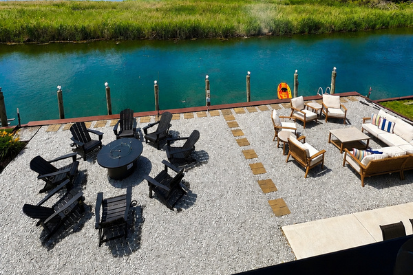 Wide waterfront patio with two seating areas Adirondack chairs around a fire pit and a lounge set overlooking calm canal water.