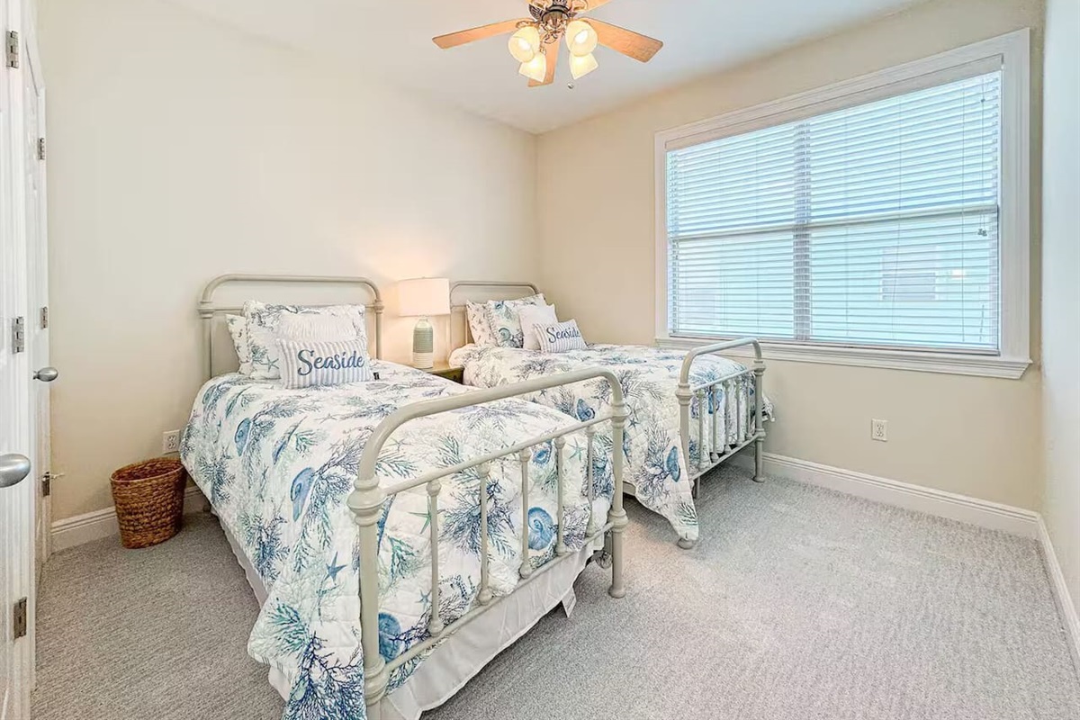 Bright guest bedroom with two twin beds, coastal-themed linens, and plenty of natural light. Includes a ceiling fan and cozy setup—perfect for kids or friends.