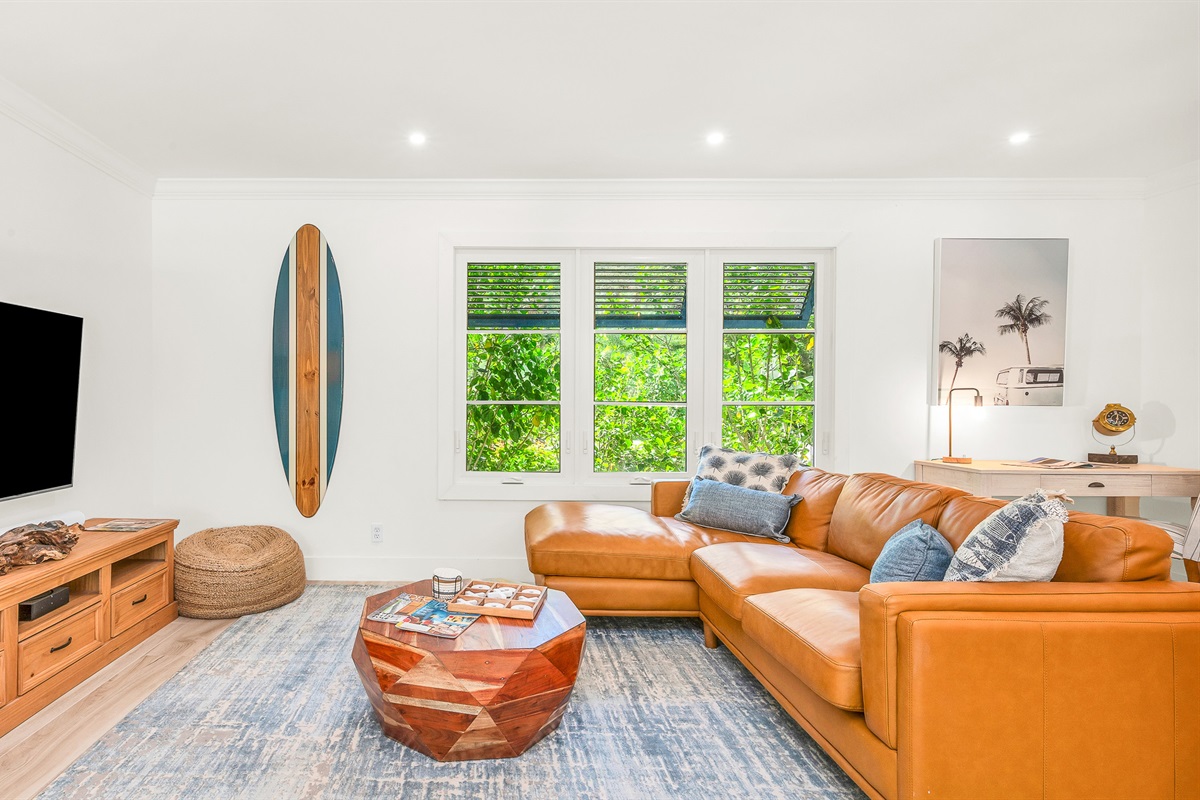 The bright and airy living room features a modern leather sectional, warm wood accents, and lush greenery views through large picture windows. Stylish touches like the surfboard décor and geometric coffee table create a relaxed coastal vibe.