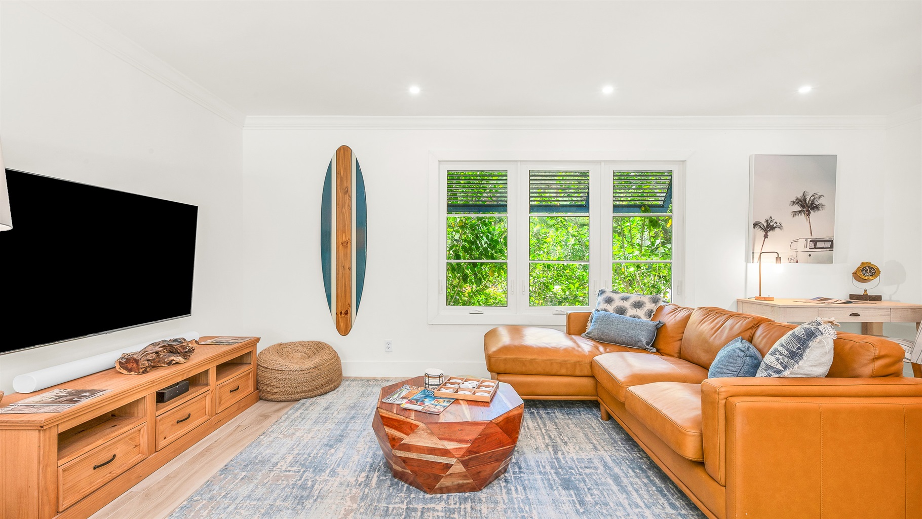 The bright and airy living room features a modern leather sectional, warm wood accents, and lush greenery views through large picture windows. Stylish touches like the surfboard décor and geometric coffee table create a relaxed coastal vibe.