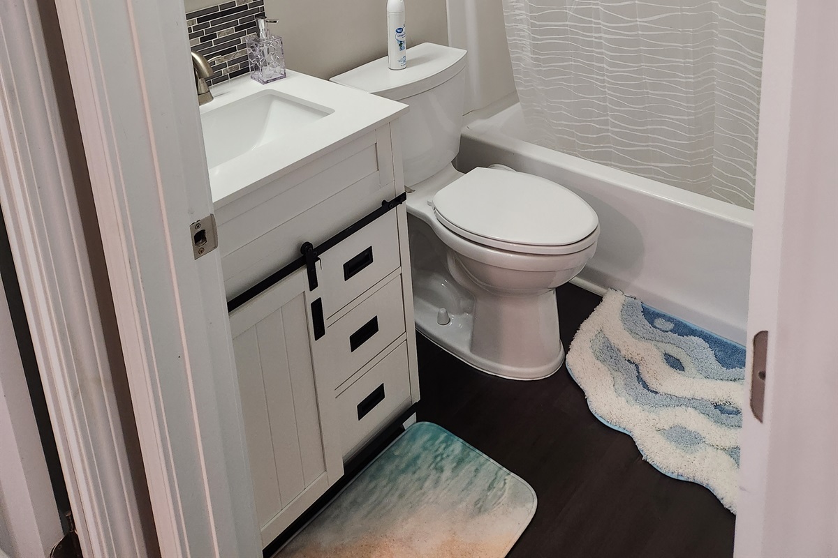 This compact bathroom is packed with style and convenience, featuring a sleek white vanity with sliding barn-style hardware, a full tub and shower combo, and playful beach-themed rugs that add a splash of coastal charm.