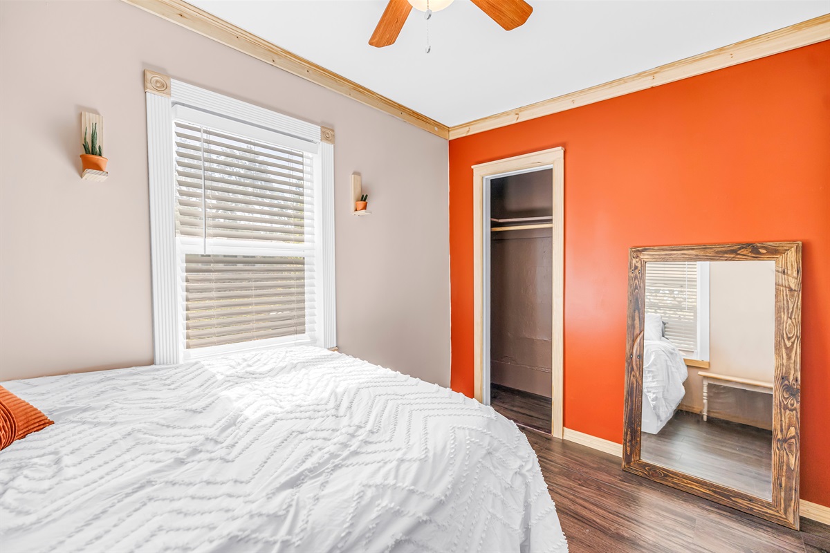 Bright bedroom with an orange accent wall, natural light, ceiling fan, and a full-length mirror for added convenience.