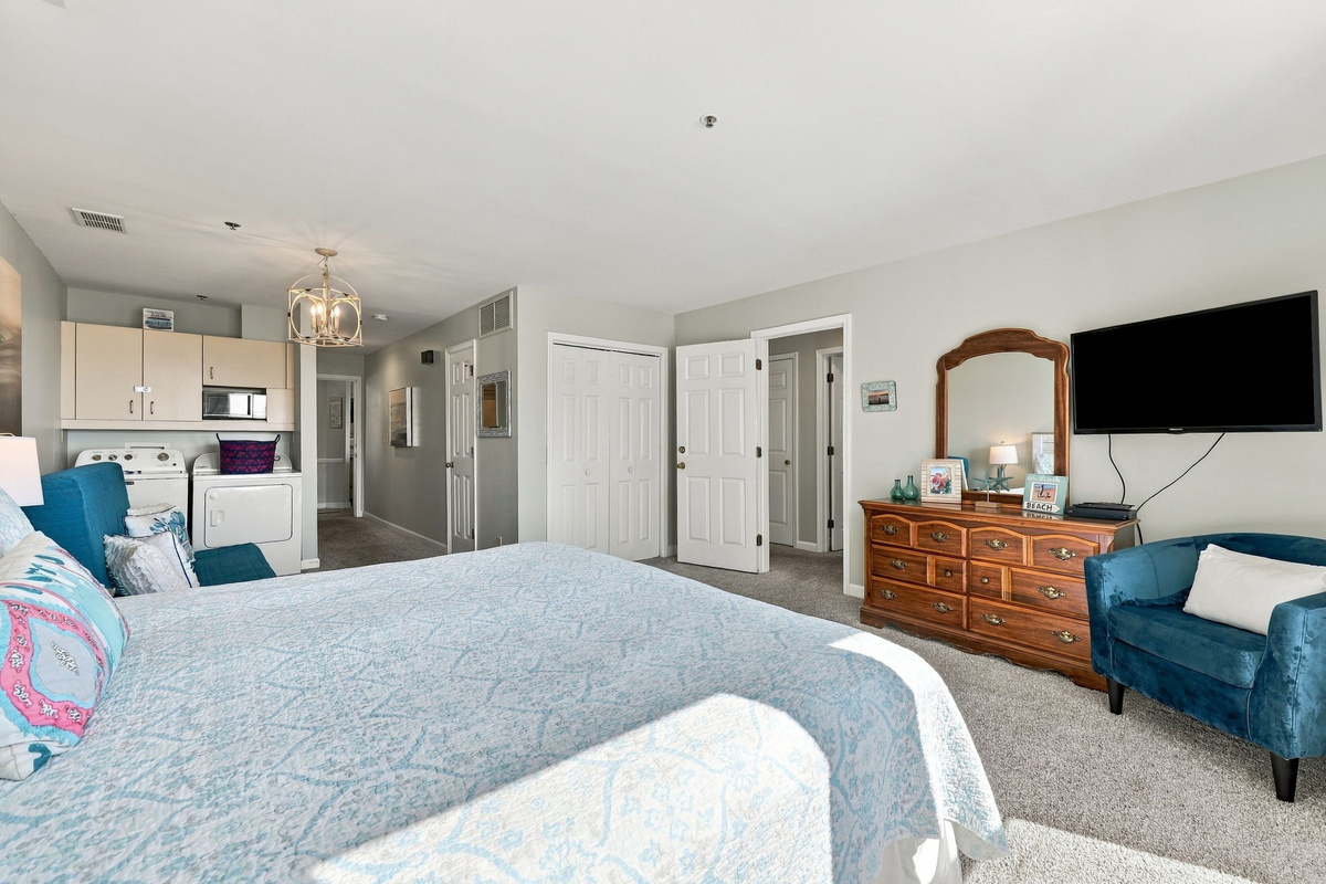 Primary suite layout showing king bed, traditional wooden dresser with mirror, wall-mounted TV, plush velvet armchair, and convenient laundry alcove in the background.   