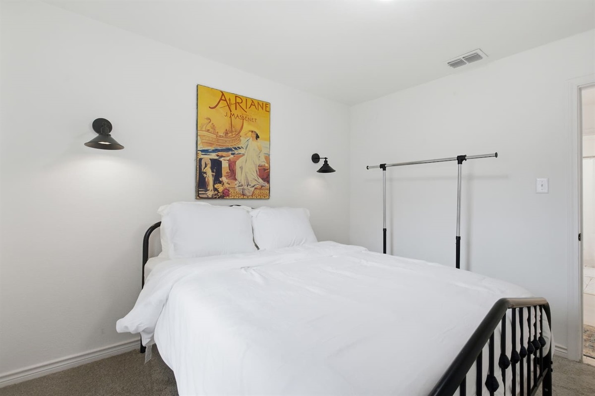 Queen bedroom with fresh linens, modern wall lighting, clothing rack, and room darkening shades for restful sleep.
