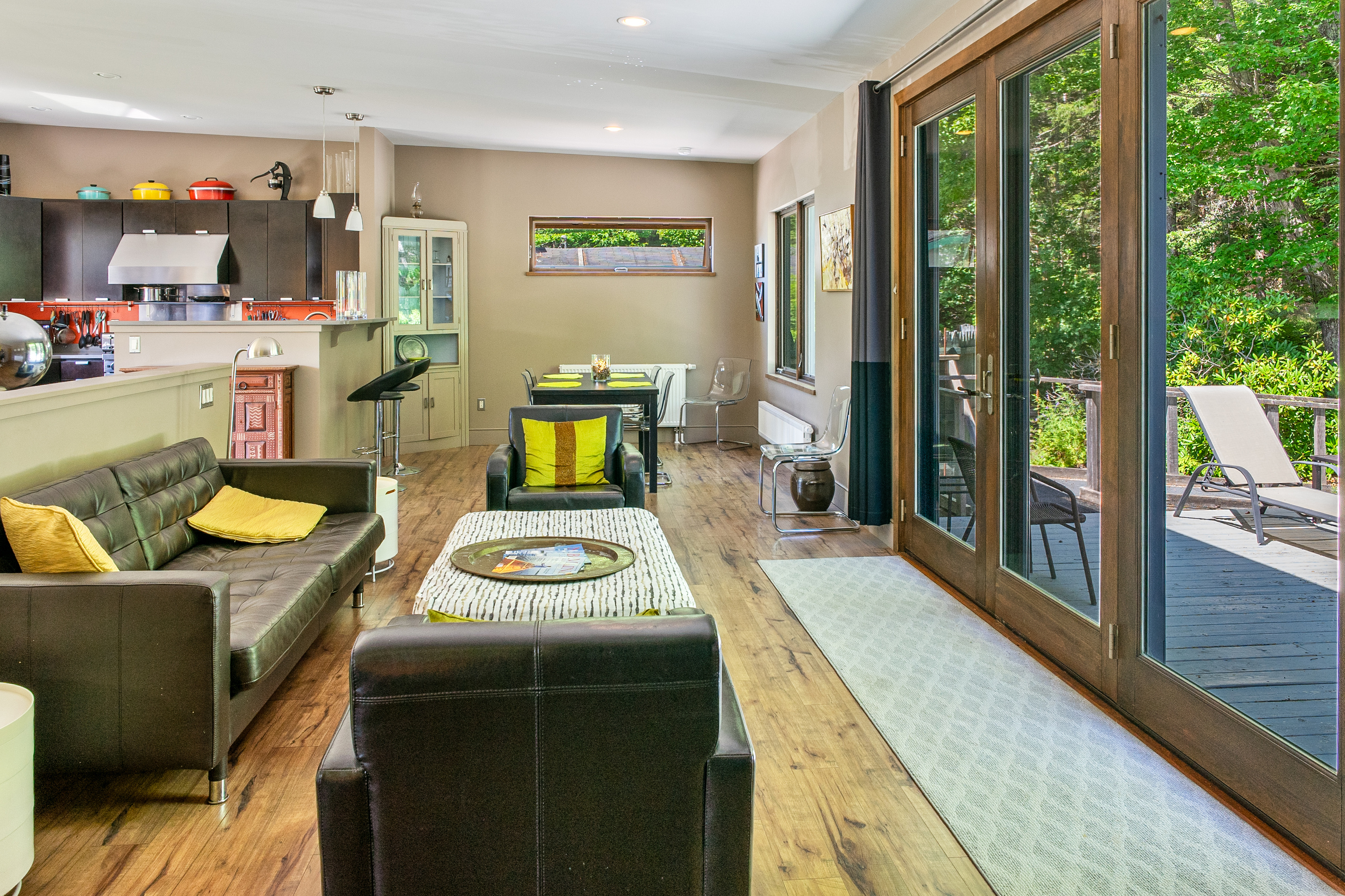 The Social Heart of "The Crag." Tufted leather seating, rich hardwood floors, and expansive floor-to-ceiling glass doors connect the open living room directly to the outdoor deck and private Acadia National Park backdrop.