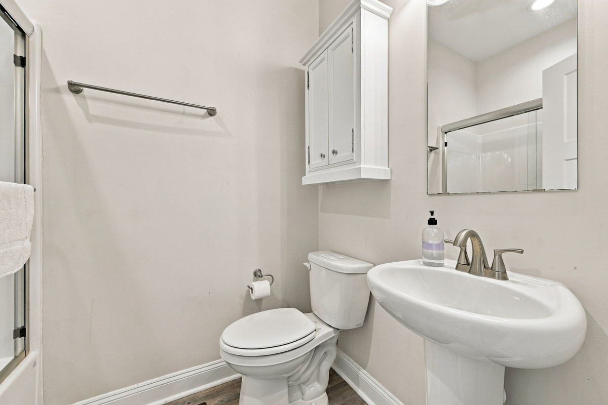 Alternate angle of the bright guest bathroom showing the white pedestal sink with brushed nickel hardware, a wall-mounted mirrored medicine cabinet, neutral painted walls, and glass-doored shower enclosure access.   