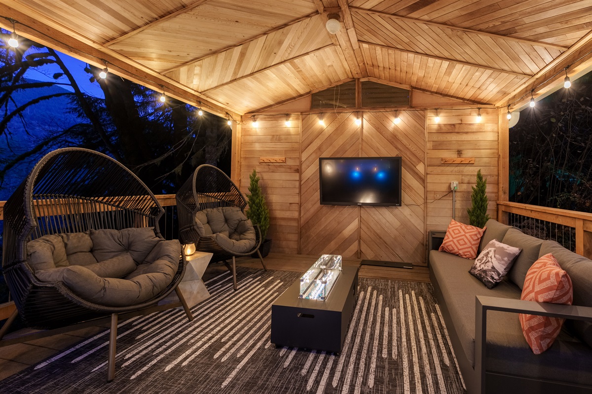 Unwind in the cozy covered lounge, complete with plush seating, warm fire feature, string lights, and an outdoor TV—your perfect nighttime hangout surrounded by the forest.