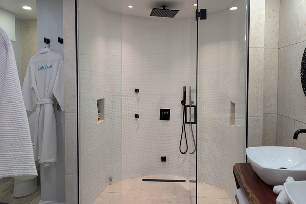 Glass-enclosed primary steam shower at Villa Coral featuring a 12-inch matte black rainfall showerhead, body jets, a handheld sprayer, white penny tile, and custom embroidered luxury robes in Dorado, Puerto Rico
