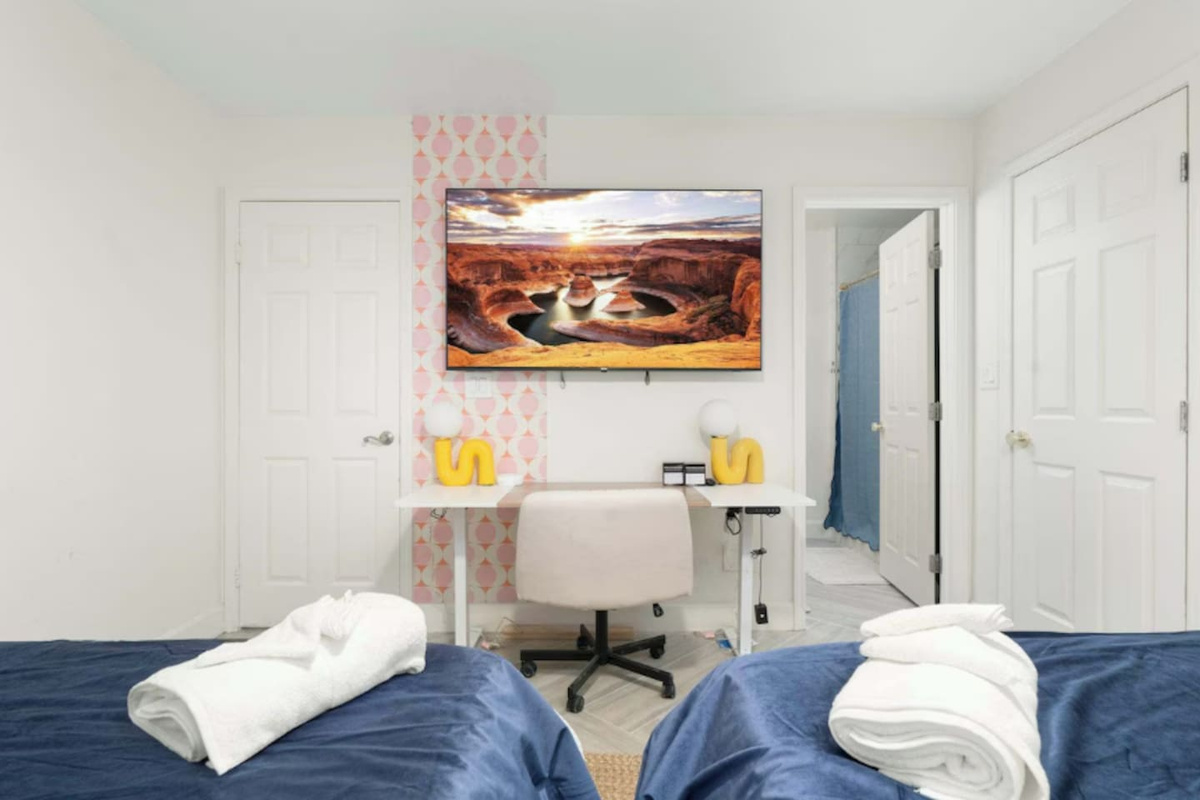 Guest bedroom with dual beds, pink wallpaper, desk, and desert landscape art—comfortable space for couples or shared stays.
