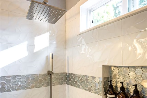 - Sleek shower with rainfall head, handheld sprayer & contemporary hardware finishes
- White textured tiles accented with metallic hexagonal band for a designer look
- Built in recessed shelf keeps bath products organized & within easy reach for use