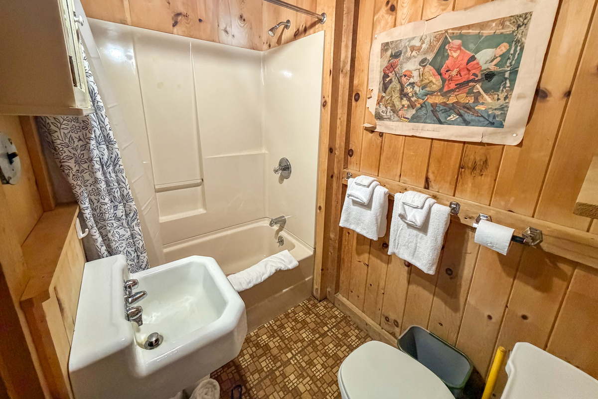 Full bathroom at Loonrise Point! Features a shower/tub combo perfect for bathing kids after a lake day. Complete with fresh towels, sink, and knotty pine walls with vintage camp art. Convenient setup on Beech Hill Pond in Otis, ME.