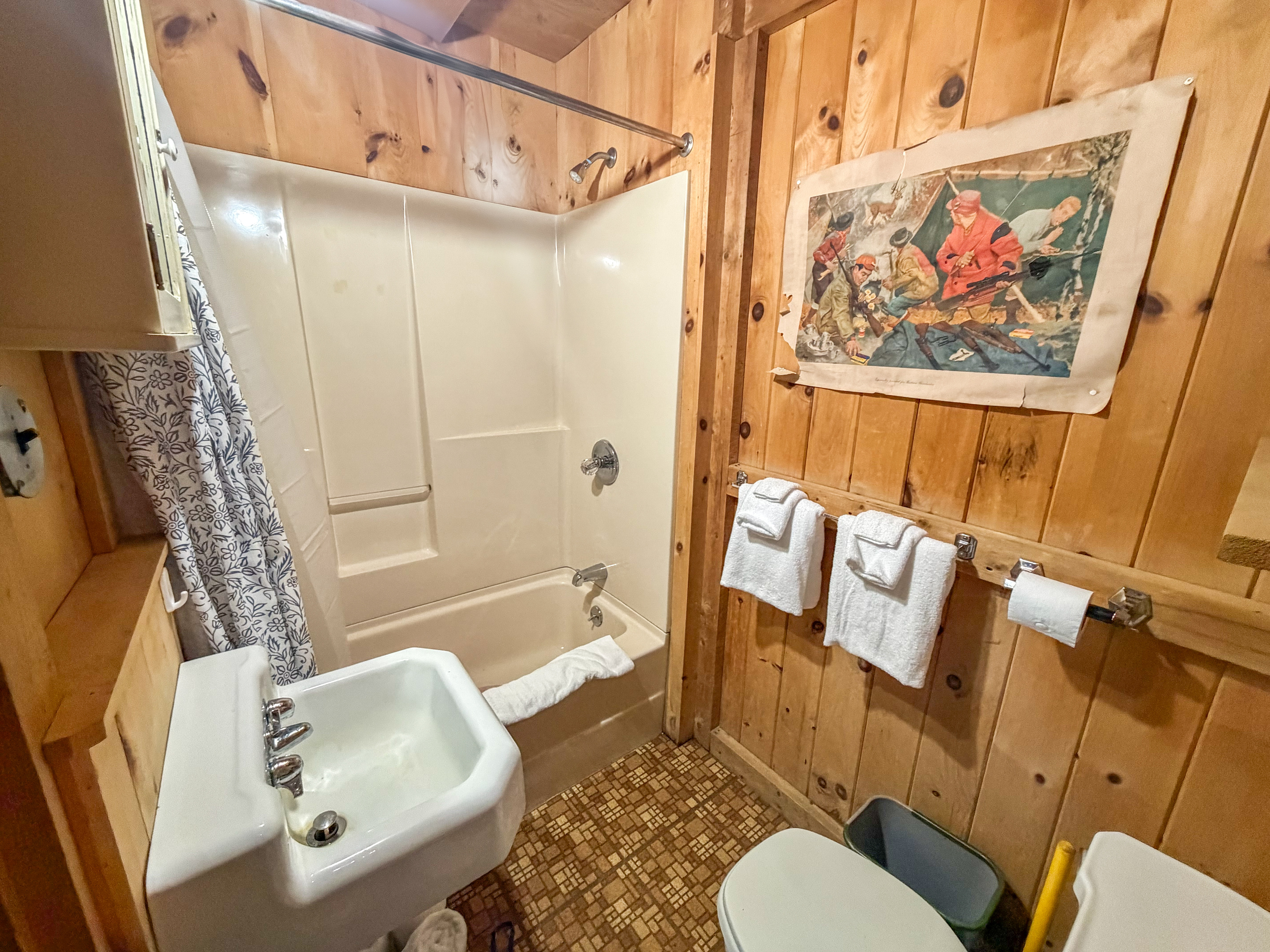 Full bathroom at Loonrise Point! Features a shower/tub combo perfect for bathing kids after a lake day. Complete with fresh towels, sink, and knotty pine walls with vintage camp art. Convenient setup on Beech Hill Pond in Otis, ME.