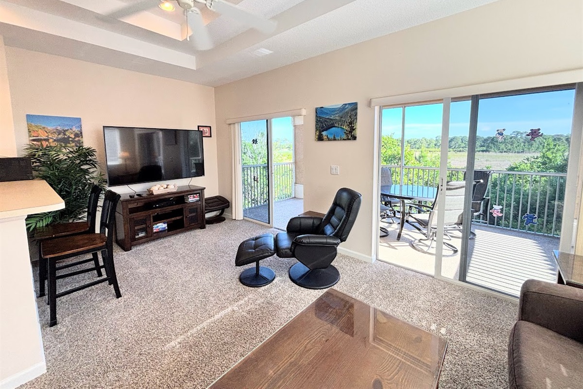 Bright living area with large TV, cozy seating, and sliding glass doors leading to the private screened balcony.