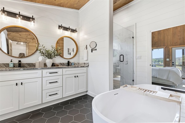 Luxurious full bathroom with freestanding soaking tub, large walk-in shower, and private water closet. White shaker vanities with ample storage, stone counters, large mirrors and warm wood accents create a serene spa-like retreat.