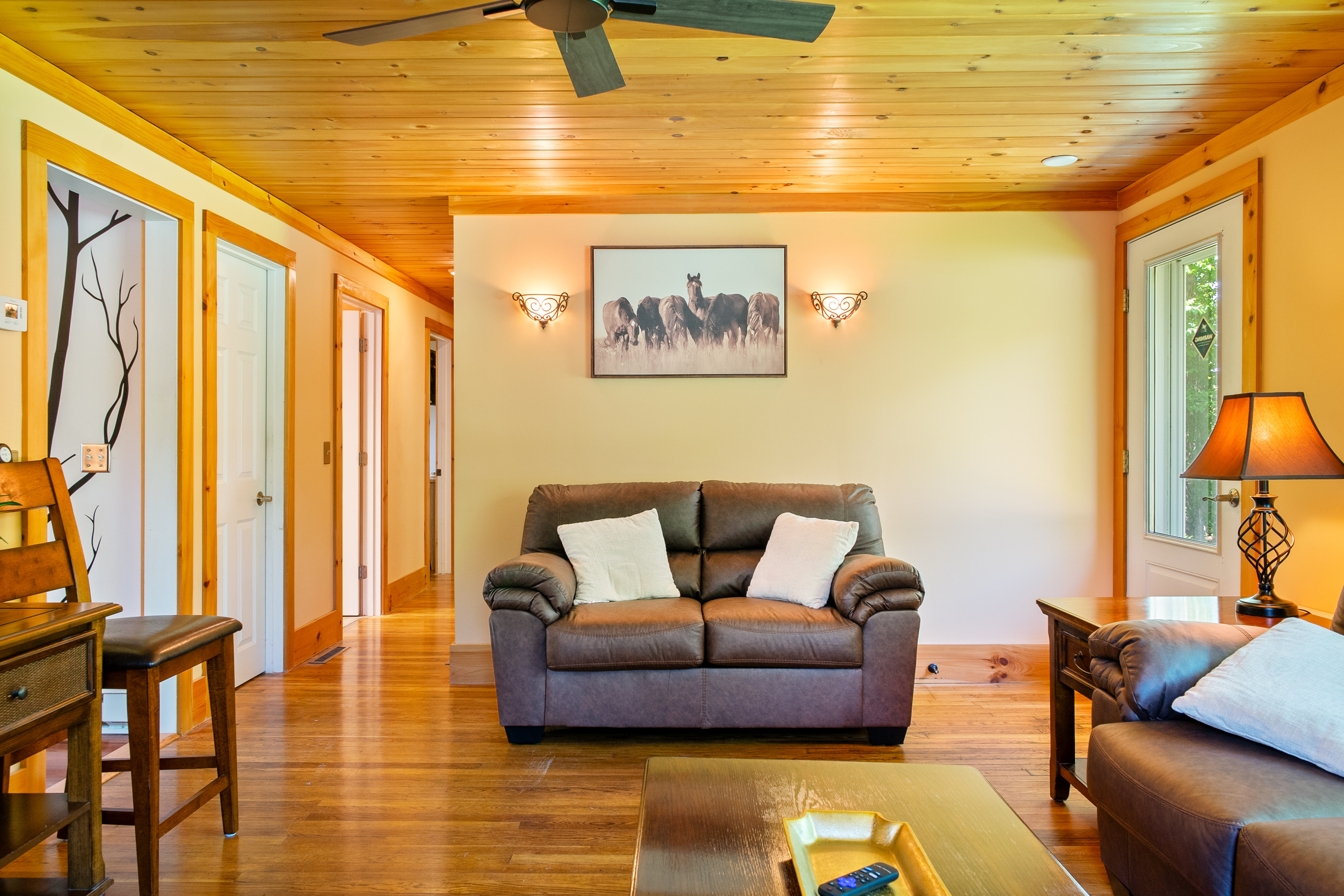 View the complete living room layout revealing the two-cushion loveseat centered on the back wall. This perspective showcases the seamless flow to the side exit door, the main couch area, and the open hallway leading to the private bedrooms.