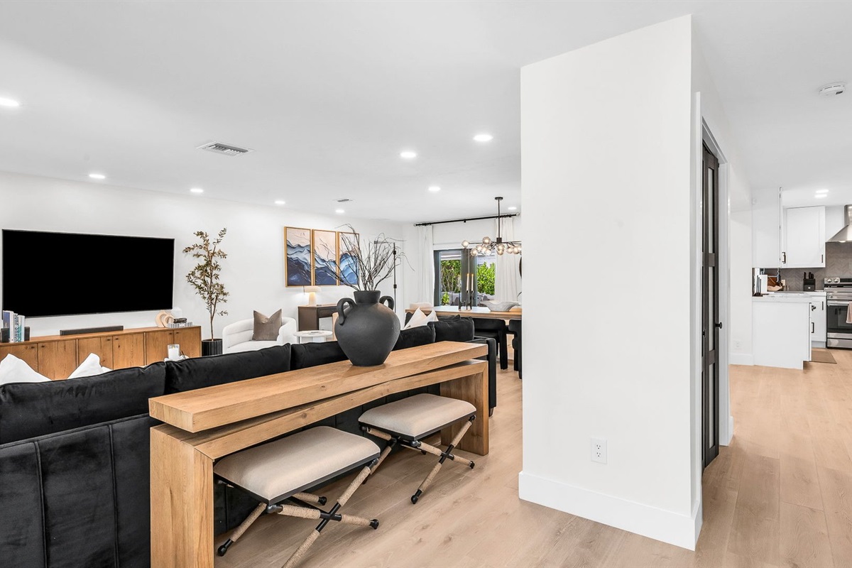 Elegant open-concept living room featuring a plush black velvet sectional, 75” smart TV, and designer mid-century modern furnishings. Floor-to-ceiling windows and French doors fill the space with natural light and connect seamlessly to the outdoor pool.