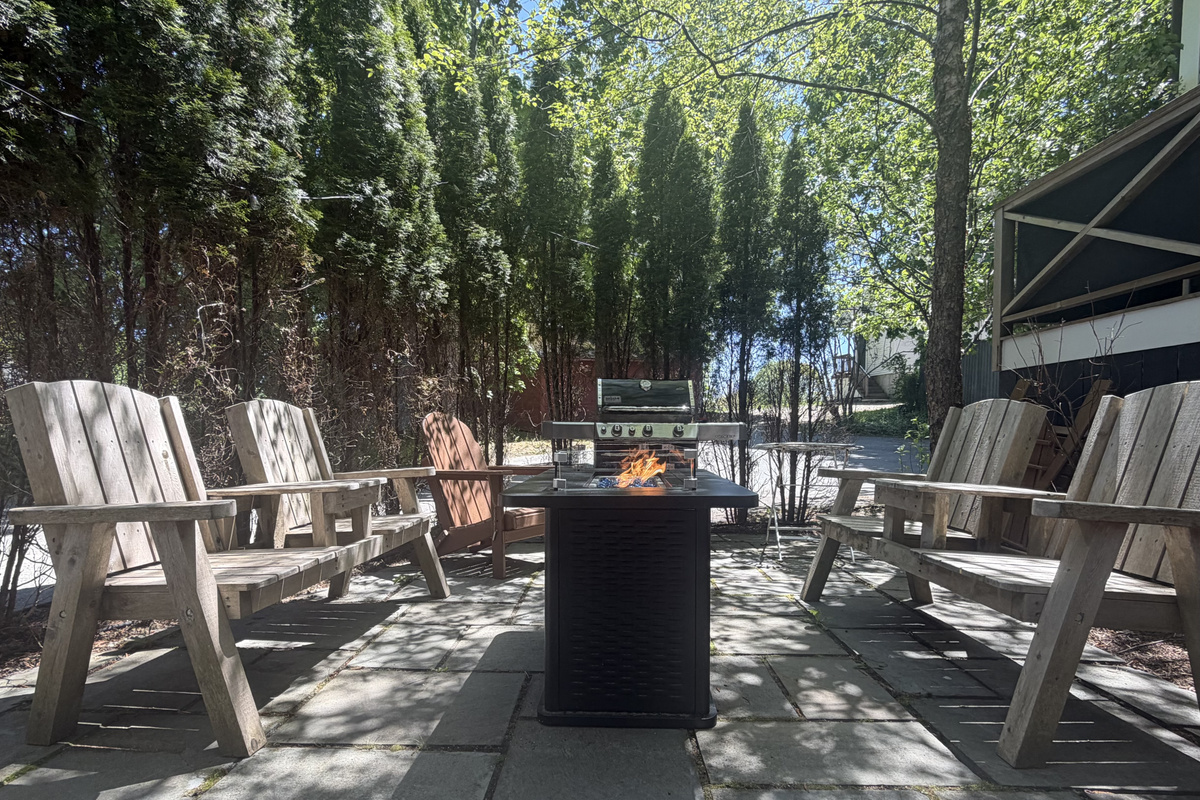 Symmetrical view of the stone patio showing the gas fire table, BBQ grill, and composite Adirondack seating. This angle highlights the arborvitae tree privacy wall that screens the outdoor gathering space from the surrounding village.