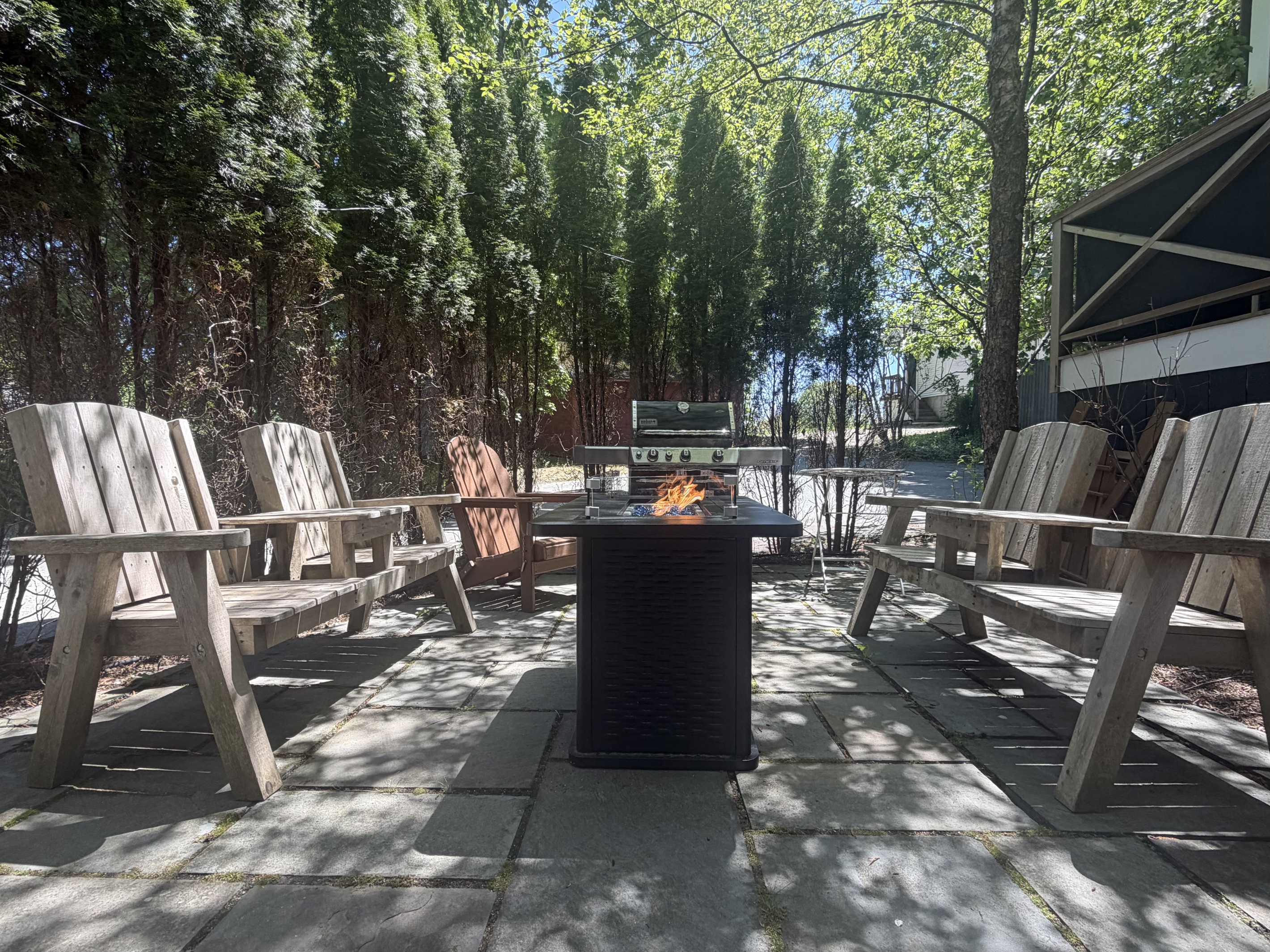 Symmetrical view of the stone patio showing the gas fire table, BBQ grill, and composite Adirondack seating. This angle highlights the arborvitae tree privacy wall that screens the outdoor gathering space from the surrounding village.