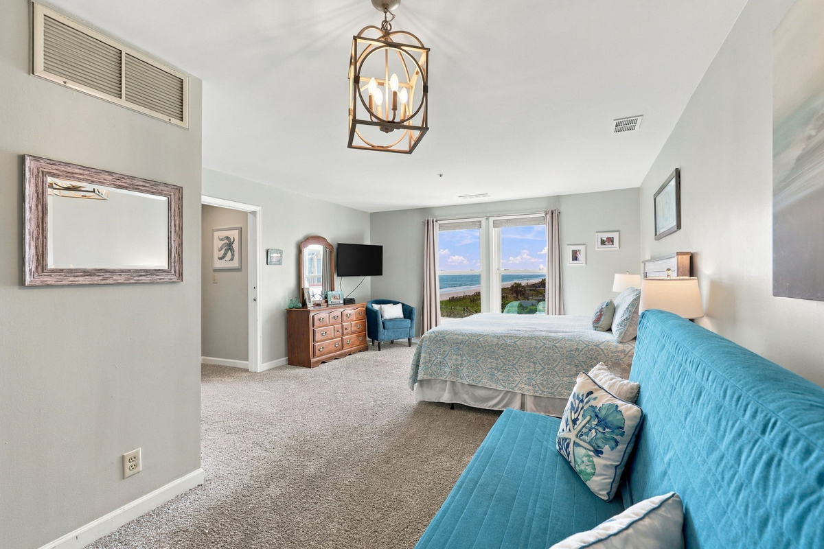Spacious oceanfront bedroom suite featuring a vibrant blue futon lounge, double bed, cage-style lantern pendant light, and direct ocean views framed by large windows.