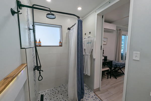Walk-in guest shower at Villa Coral featuring a matte black rainfall showerhead, pebble tile flooring, custom tilework, and complimentary luxury robes in Dorado, Puerto Rico.