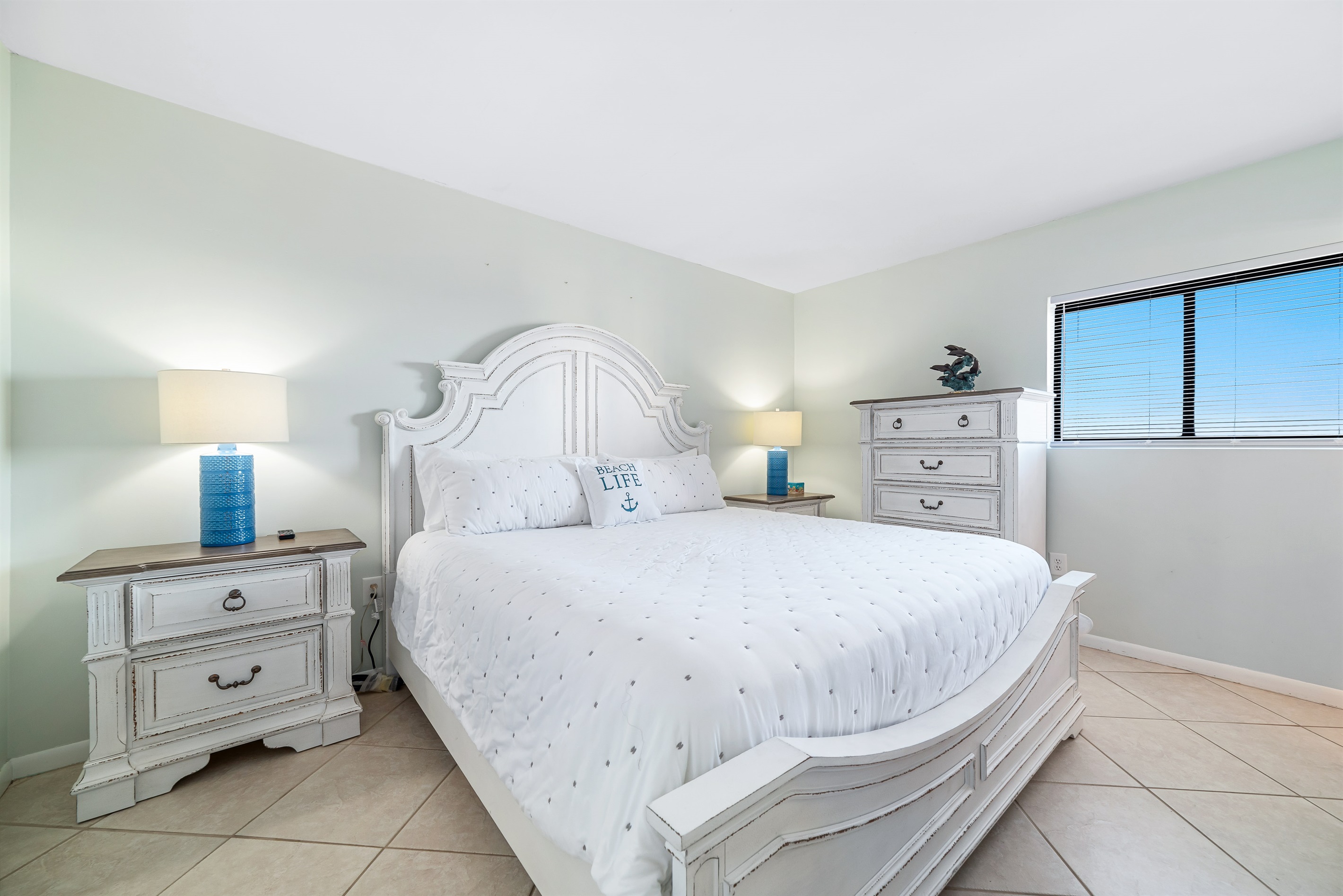 This well-appointed bedroom features a king-size bed with a statement headboard, crisp white bedding, matching nightstands with lamps, a dresser, and a window offering natural light.