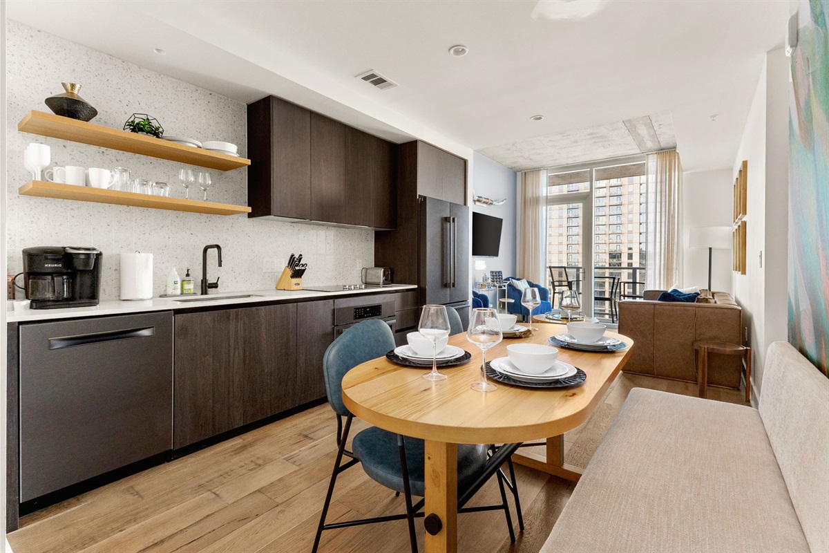 A contemporary open-concept space featuring a fully equipped kitchen with dark wood cabinetry and open shelving, which flows into a stylish dining area with an oval wooden table and built-in bench, and a comfortable living room with a balcony offering a c
