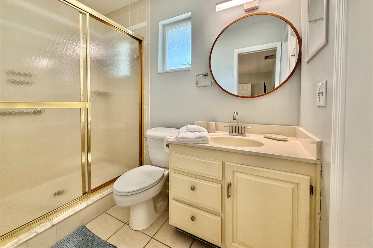 This clean and cozy bathroom features a spacious walk-in shower with gold trim, a round mirror, and natural light from the frosted window.