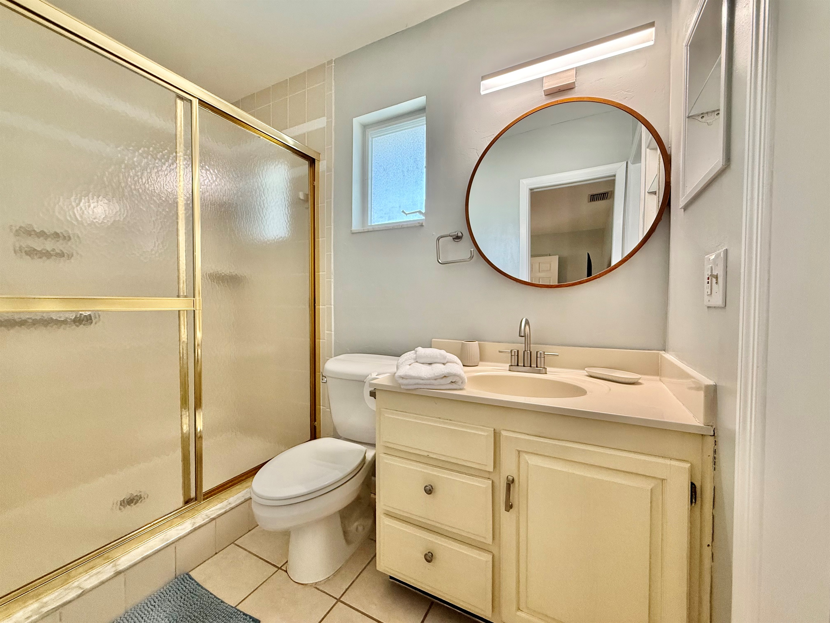 This clean and cozy bathroom features a spacious walk-in shower with gold trim, a round mirror, and natural light from the frosted window.