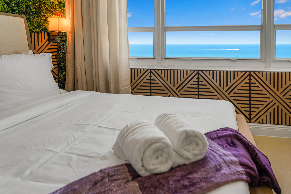 Fresh rolled towels and soft throws await on arrival, with the ocean just beyond the window behind the bed.