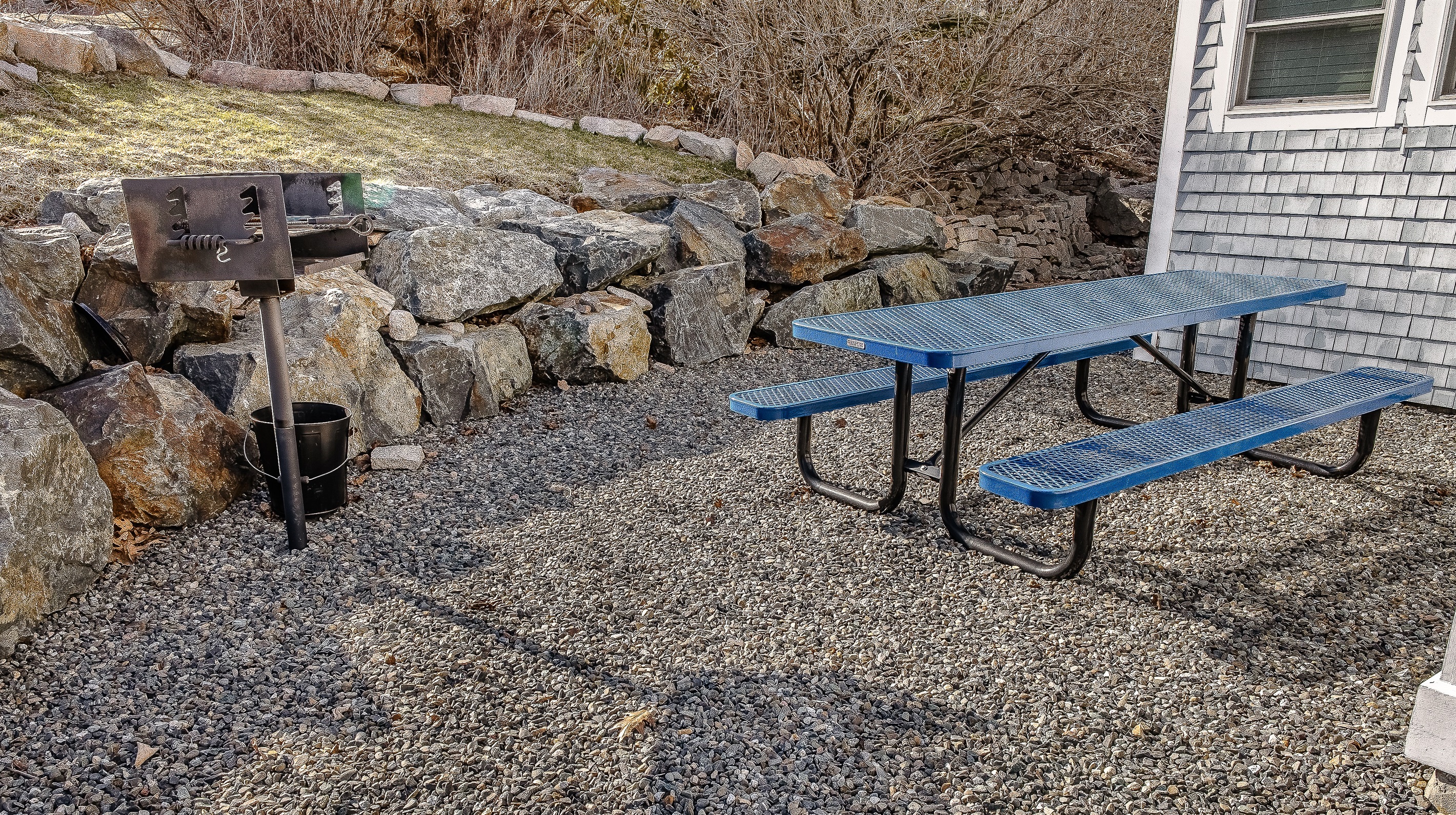 A classic picnic table is ready for your group, nestled against a retaining wall of genuine Maine granite. This dedicated outdoor space, featuring a built-in charcoal grill, provides a relaxed, authentic atmosphere for refuel and recovery after your hike.