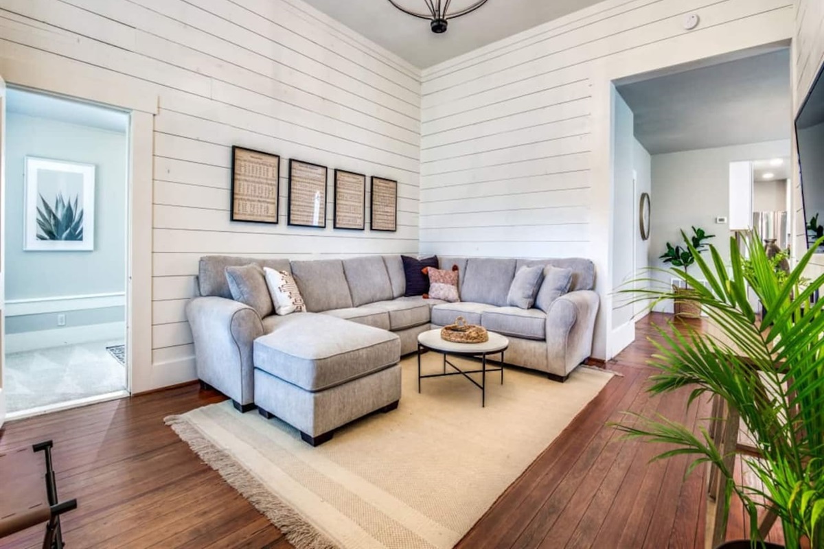 Corner view of the bright living room with full sectional, woven rug, hardwood floors, chandelier, and dual windows. Dedicated workspace available. Modern farmhouse Airbnb near Denton County wedding venues in downtown Aubrey TX.