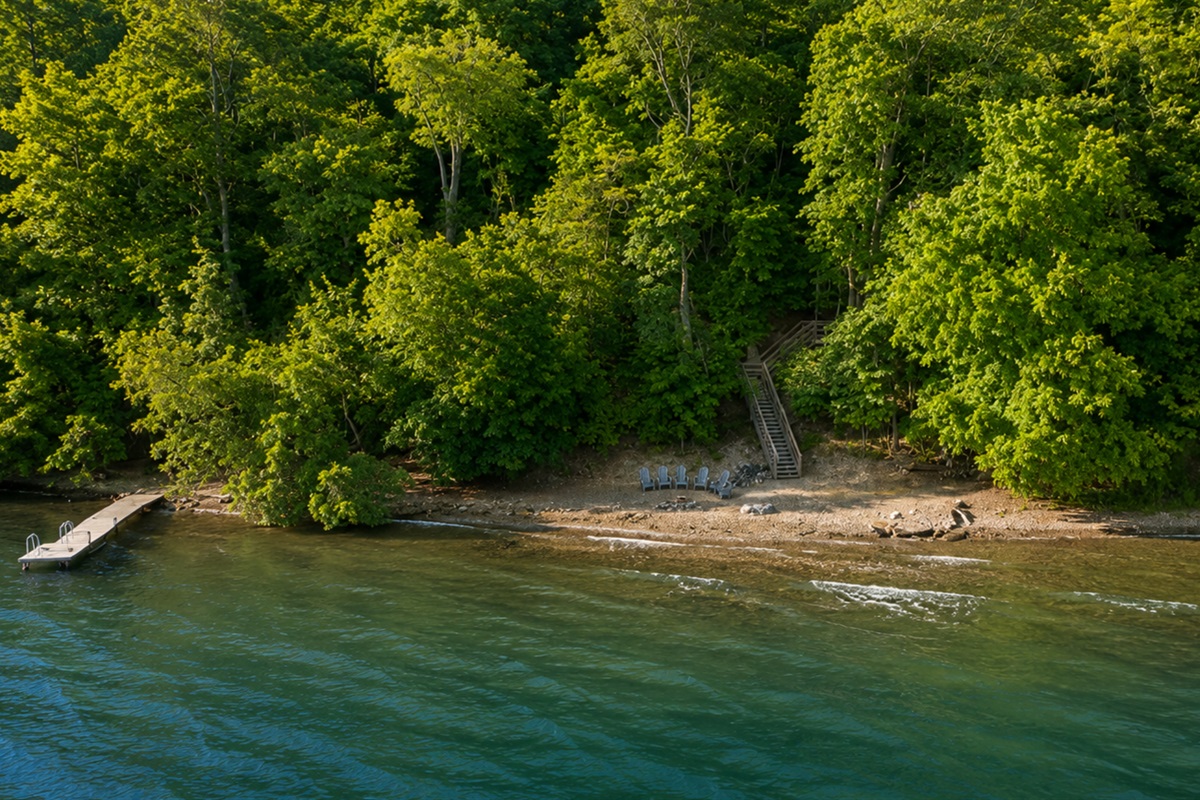 Aerial view of a private, wooded shoreline retreat with direct lake access, dock, and Adirondack chairs for lakeside relaxation.