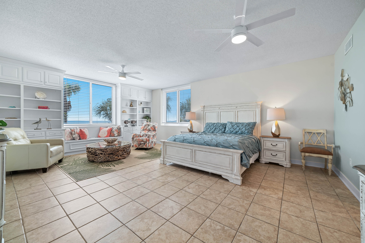 This tranquil primary bedroom is bathed in natural light, showcasing built-in shelving, a comfortable seating area, and stunning views.