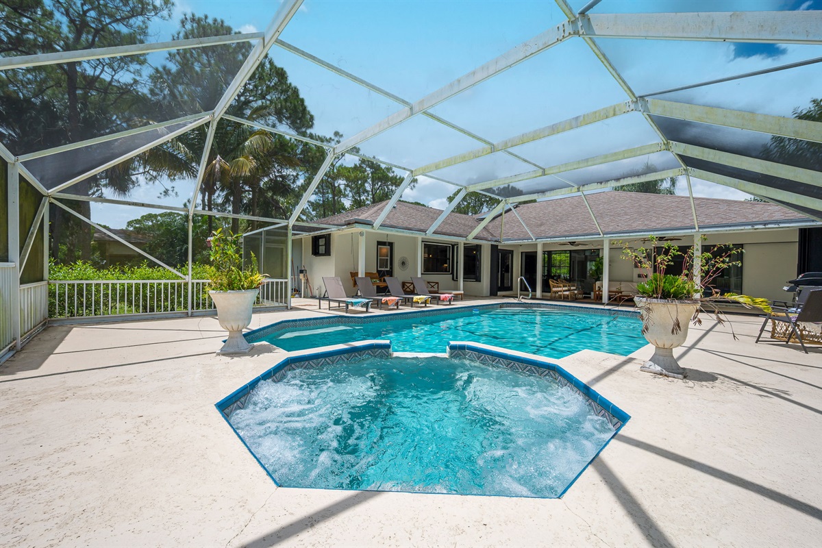Screened-in pool area with heated pool and spa, surrounded by lush greenery. Features ample lounge seating, BBQ grill, and cozy covered patio with outdoor dining and relaxing sitting area.
