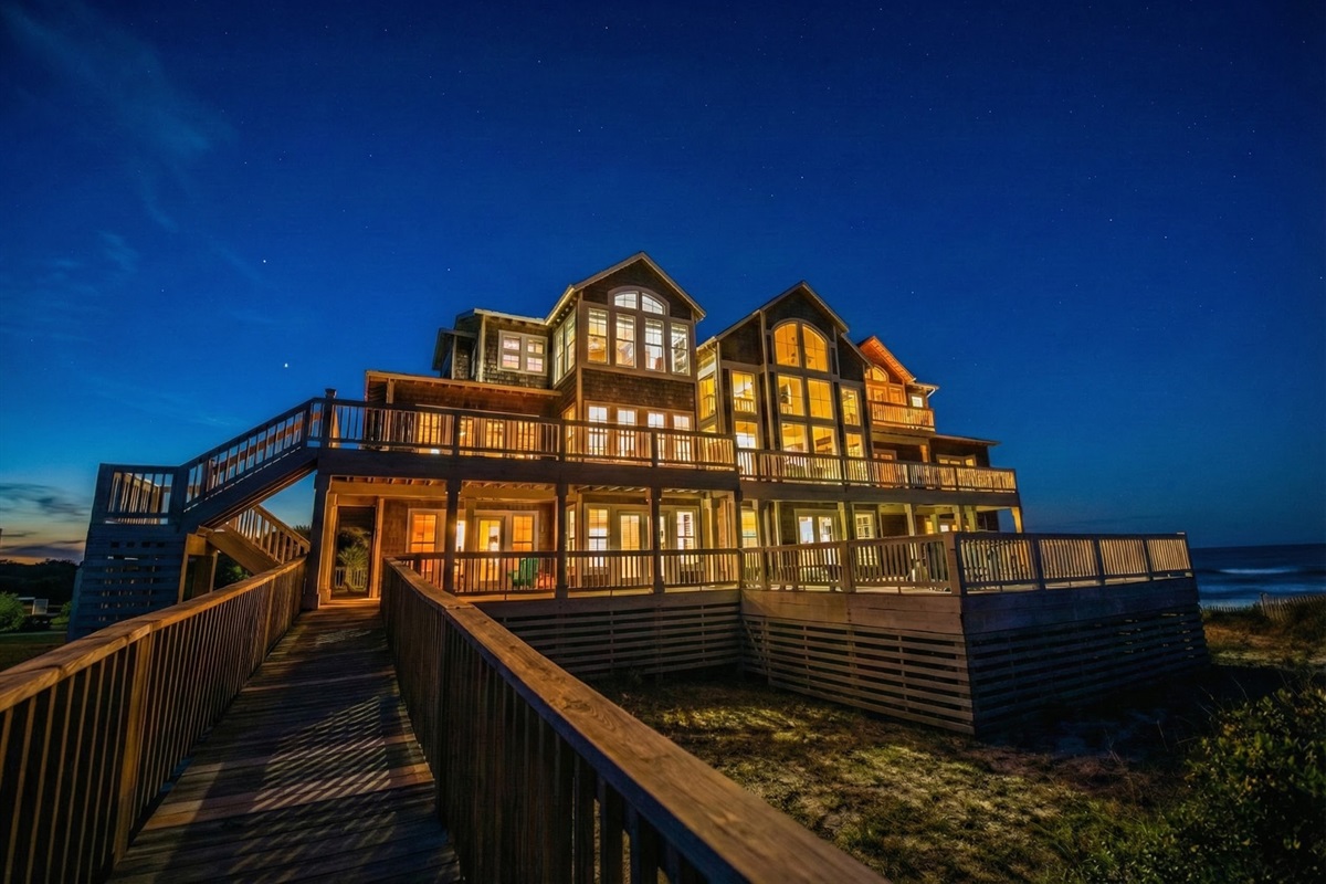 A twilight view highlighting the grandeur of the home, featuring multi-level wrap-around decks and a private wooden boardwalk leading directly to the beach.