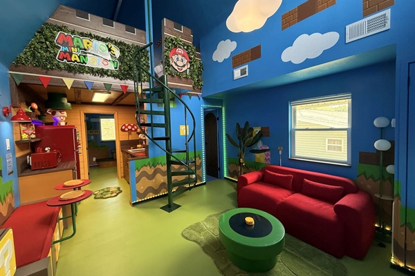 Explore the "castle" area with spiral staircase to the lofted Mario's Mini Mansion king bed! This vibrant blue living room features a squishy red sofa, a green coffee table, custom signage, and playful Mario details -- endless adventures for all.